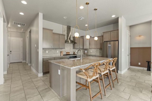 kitchen featuring a center island with sink, high quality fridge, a kitchen bar, decorative light fixtures, and wall chimney exhaust hood