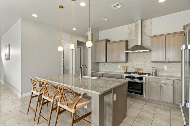 kitchen featuring wall chimney exhaust hood, sink, decorative light fixtures, stainless steel appliances, and a kitchen island with sink