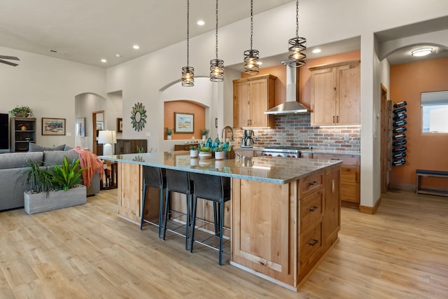 kitchen with light stone counters, tasteful backsplash, light wood-type flooring, light brown cabinets, and wall chimney range hood