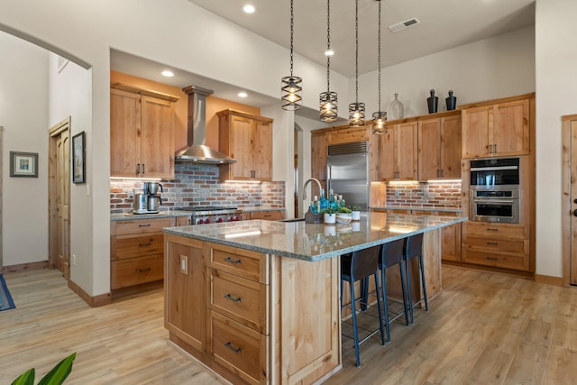 kitchen featuring appliances with stainless steel finishes, stone countertops, pendant lighting, a kitchen island with sink, and wall chimney exhaust hood