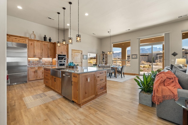 kitchen with pendant lighting, light hardwood / wood-style flooring, a kitchen island with sink, stainless steel appliances, and light stone counters