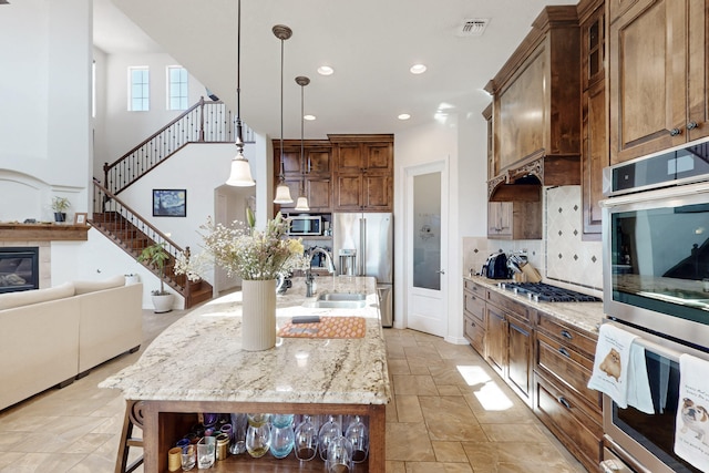 kitchen with appliances with stainless steel finishes, tasteful backsplash, light stone counters, an island with sink, and decorative light fixtures