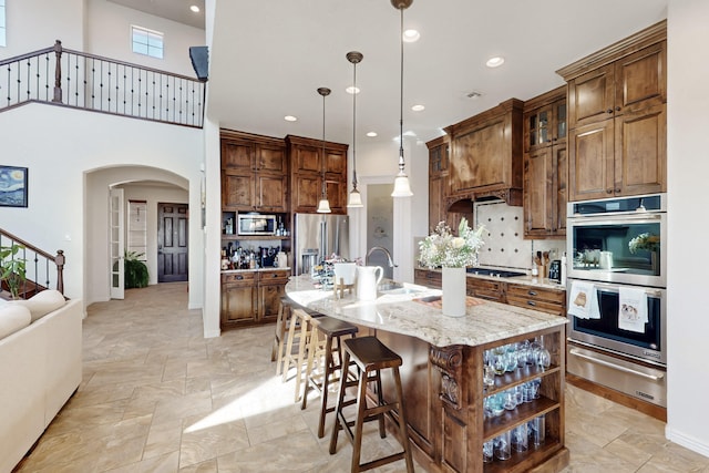 kitchen featuring sink, hanging light fixtures, light stone counters, stainless steel appliances, and a center island with sink