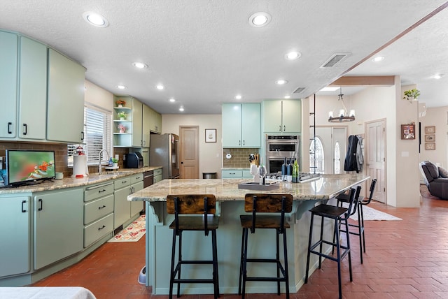 kitchen featuring sink, green cabinets, appliances with stainless steel finishes, backsplash, and a center island with sink