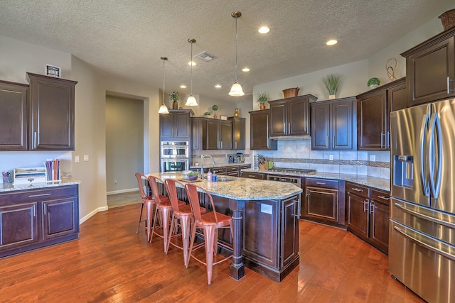 kitchen with appliances with stainless steel finishes, a kitchen bar, hanging light fixtures, a kitchen island with sink, and light stone countertops