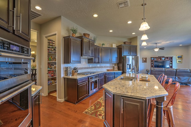 kitchen with decorative light fixtures, sink, dark brown cabinetry, stainless steel appliances, and a center island with sink