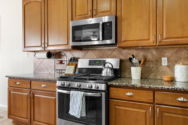 kitchen with brown cabinets, backsplash, stainless steel appliances, and dark stone countertops