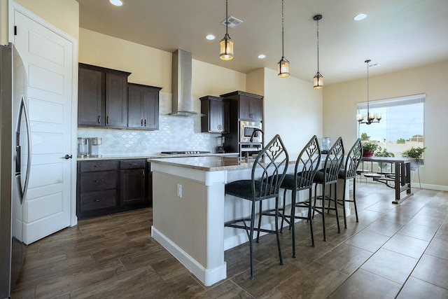 kitchen featuring a kitchen breakfast bar, wall chimney range hood, stainless steel appliances, hanging light fixtures, and a kitchen island with sink