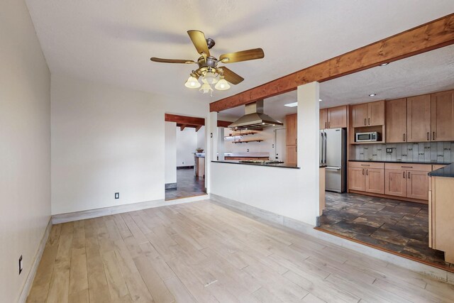 kitchen featuring wood finished floors, beam ceiling, appliances with stainless steel finishes, dark countertops, and wall chimney exhaust hood
