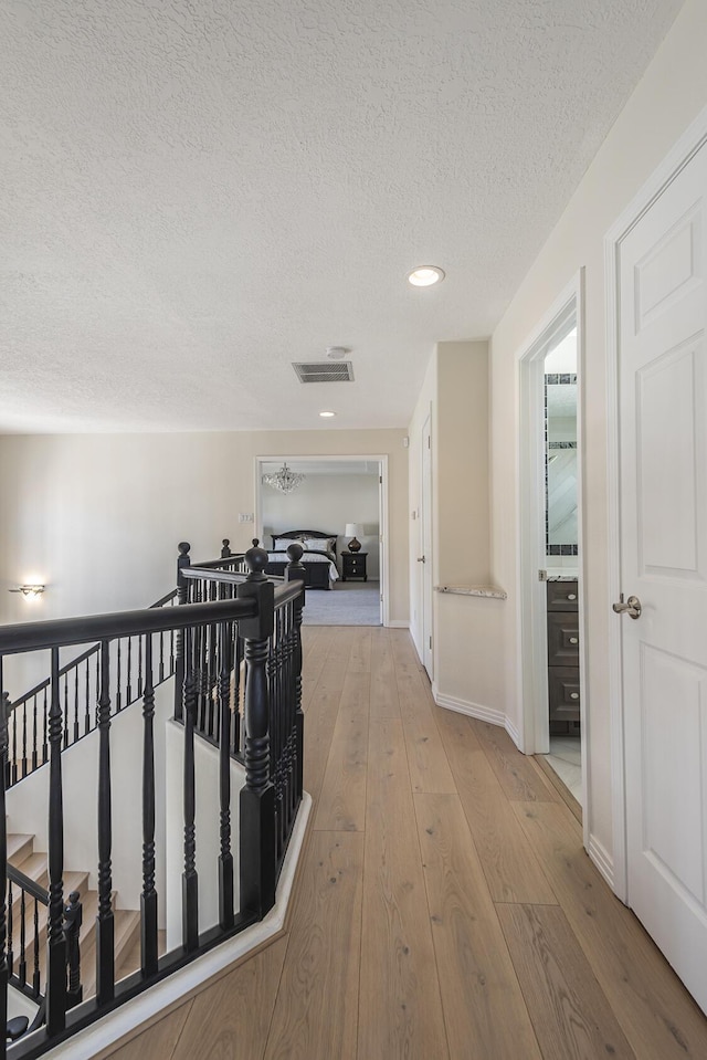 hallway with baseboards, visible vents, light wood-style flooring, a textured ceiling, and an upstairs landing