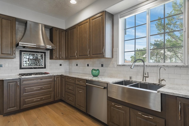 kitchen featuring light stone counters, stainless steel appliances, a sink, light wood-type flooring, and wall chimney exhaust hood