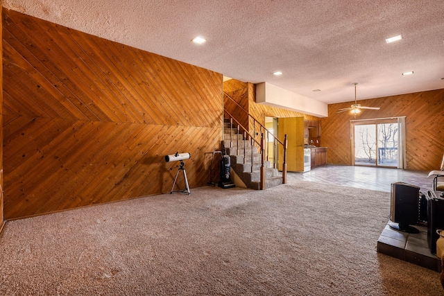 carpeted living room with stairs, wooden walls, a ceiling fan, and a textured ceiling