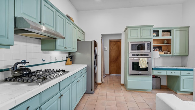 kitchen featuring under cabinet range hood, appliances with stainless steel finishes, tile counters, open shelves, and tasteful backsplash