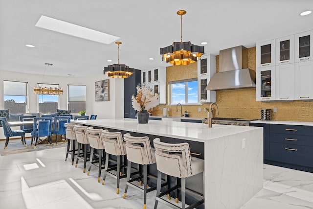 kitchen with a spacious island, hanging light fixtures, backsplash, wall chimney range hood, and white cabinets