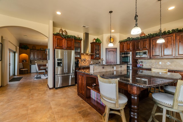 kitchen featuring arched walkways, wall chimney exhaust hood, a kitchen island with sink, stainless steel appliances, and open shelves