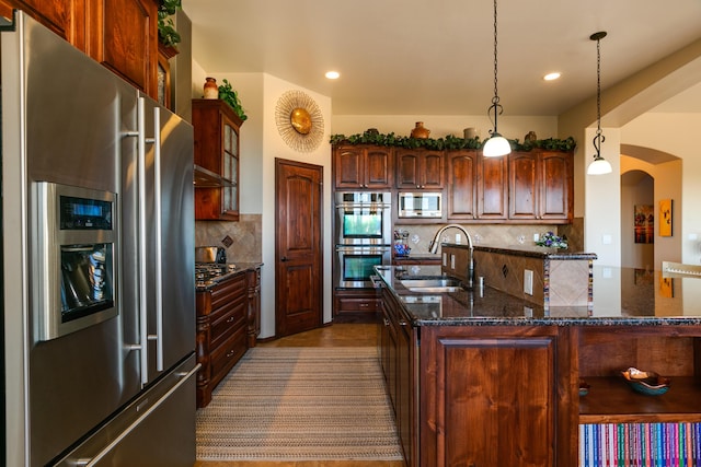 kitchen with arched walkways, stainless steel appliances, tasteful backsplash, a sink, and an island with sink