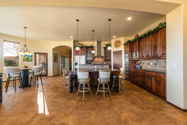 kitchen featuring arched walkways, a breakfast bar area, stainless steel appliances, a kitchen island, and wall chimney range hood