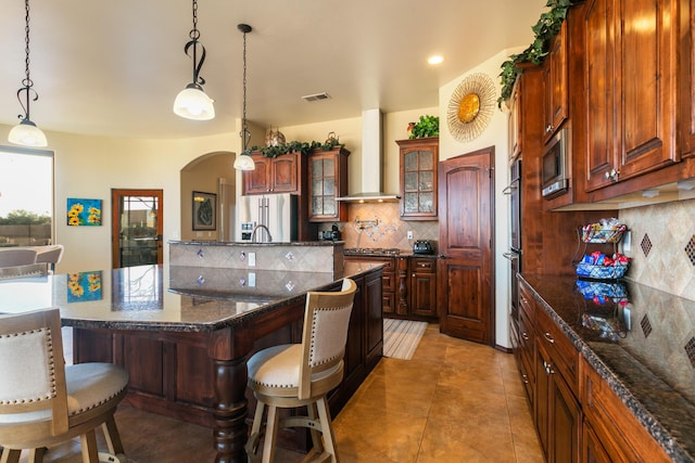 kitchen with arched walkways, stainless steel appliances, decorative backsplash, wall chimney range hood, and a kitchen bar