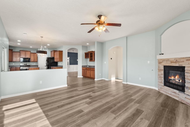 unfurnished living room with a textured ceiling, a stone fireplace, wood finished floors, a ceiling fan, and baseboards