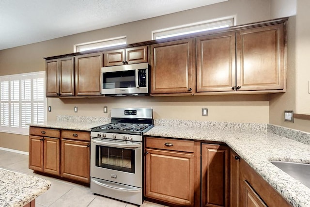 kitchen with light tile patterned floors, brown cabinetry, appliances with stainless steel finishes, light stone counters, and a sink