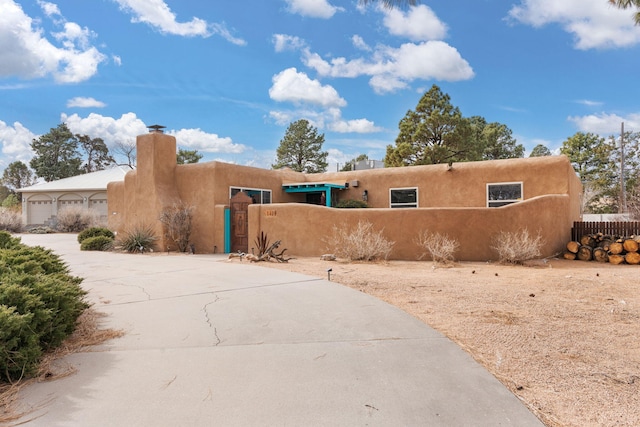 pueblo revival-style home featuring driveway, a garage, a fenced front yard, a chimney, and stucco siding