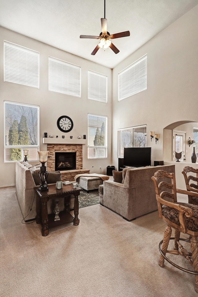 living room with arched walkways, light colored carpet, a high ceiling, a ceiling fan, and a stone fireplace