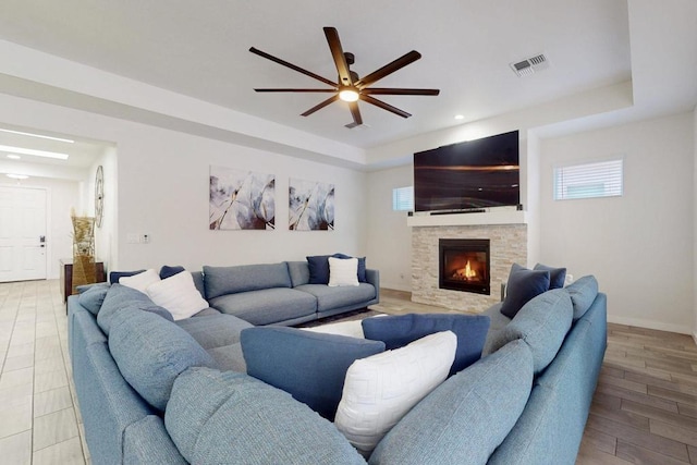 living room with light wood finished floors, a raised ceiling, visible vents, ceiling fan, and a stone fireplace