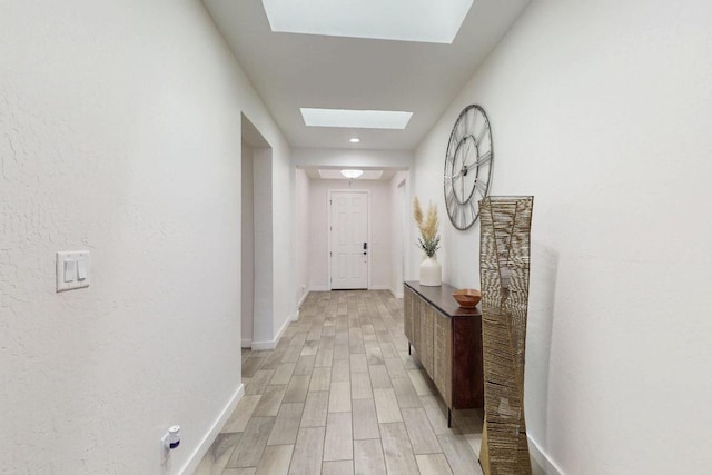 corridor featuring a skylight, baseboards, and light wood finished floors