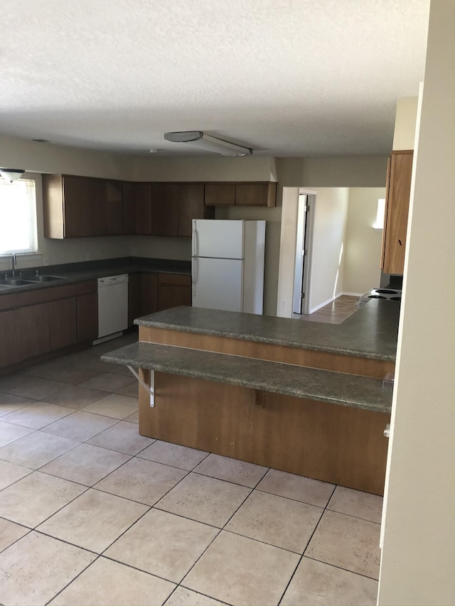 kitchen featuring light tile patterned floors, white appliances, a sink, brown cabinetry, and dark countertops