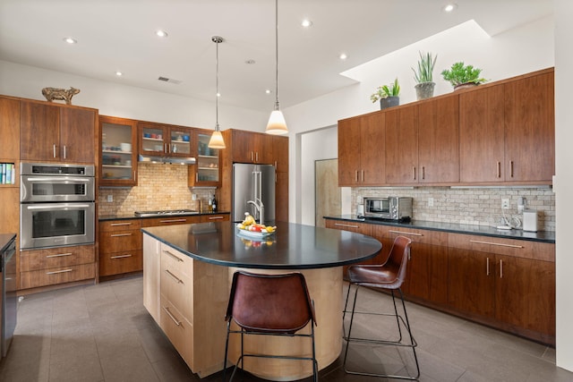 kitchen with stainless steel appliances, hanging light fixtures, a center island with sink, dark countertops, and glass insert cabinets