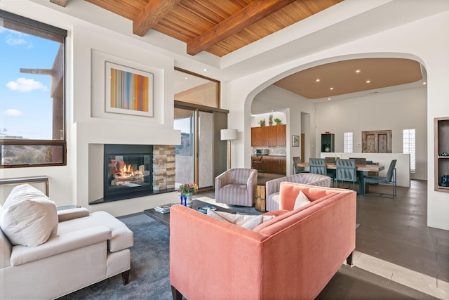 living room featuring arched walkways, wooden ceiling, a high ceiling, a stone fireplace, and beam ceiling