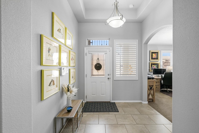 foyer featuring baseboards, arched walkways, a raised ceiling, light colored carpet, and light tile patterned flooring