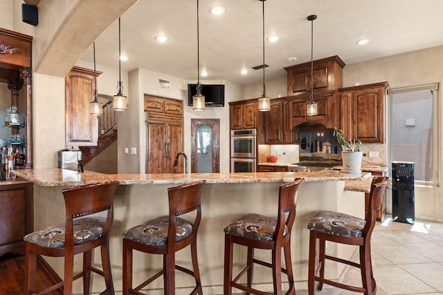 kitchen with decorative light fixtures, tasteful backsplash, double oven, light stone countertops, and a peninsula