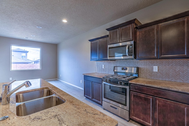 kitchen with stainless steel appliances, backsplash, a sink, dark brown cabinetry, and light stone countertops