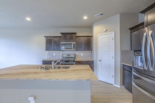 kitchen with dark brown cabinetry, a center island with sink, visible vents, stainless steel appliances, and a sink