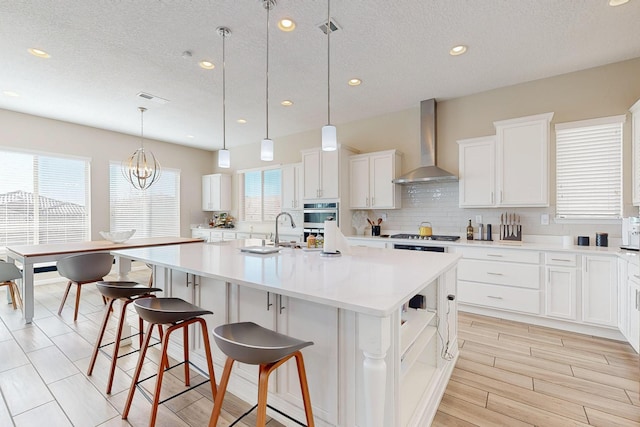 kitchen with backsplash, wall chimney range hood, light countertops, an island with sink, and white cabinets