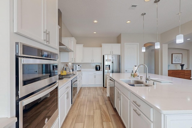 kitchen with a sink, stainless steel appliances, wall chimney range hood, decorative backsplash, and hanging light fixtures