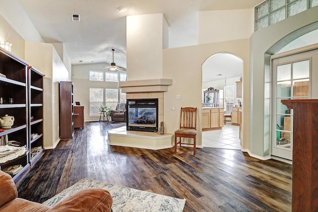 living area with arched walkways, a tile fireplace, ceiling fan, wood finished floors, and high vaulted ceiling