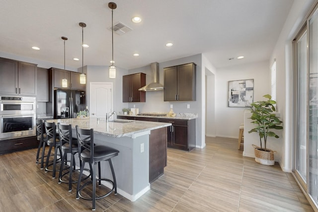 kitchen featuring dark brown cabinetry, visible vents, a breakfast bar area, stainless steel appliances, and wall chimney range hood