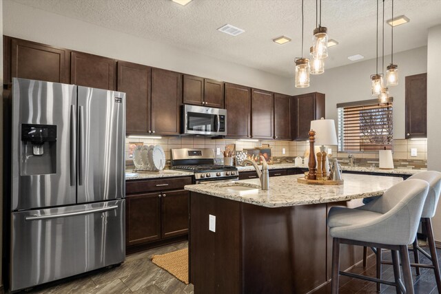 kitchen with tasteful backsplash, appliances with stainless steel finishes, a kitchen island with sink, a sink, and dark brown cabinetry