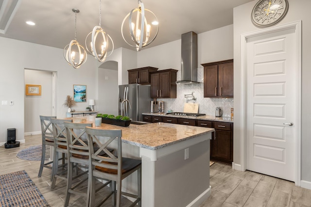 kitchen with light wood finished floors, appliances with stainless steel finishes, dark brown cabinets, wall chimney range hood, and backsplash