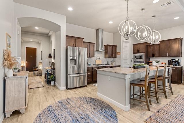 kitchen featuring light wood finished floors, stainless steel appliances, visible vents, dark brown cabinetry, and wall chimney exhaust hood