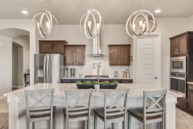 kitchen featuring light stone counters, a kitchen island with sink, light wood-style flooring, appliances with stainless steel finishes, and tasteful backsplash