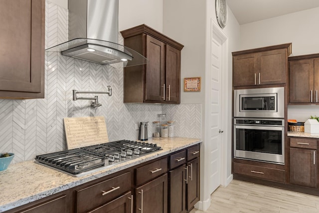 kitchen with light stone counters, stainless steel appliances, backsplash, dark brown cabinetry, and wall chimney range hood