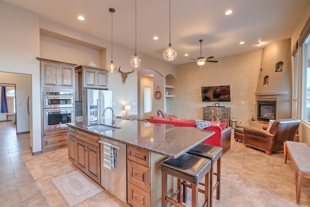 kitchen featuring arched walkways, appliances with stainless steel finishes, a glass covered fireplace, a sink, and dark stone counters