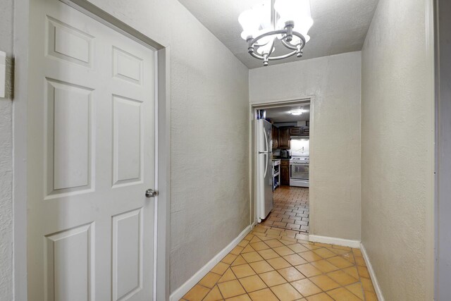 hall featuring light tile patterned flooring, a textured wall, baseboards, and an inviting chandelier