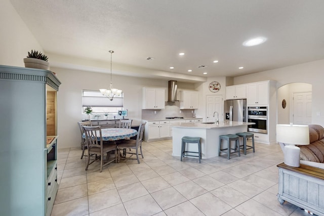 kitchen with a sink, arched walkways, appliances with stainless steel finishes, white cabinets, and wall chimney range hood