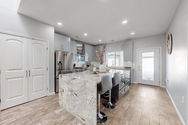 kitchen featuring wall chimney exhaust hood, appliances with stainless steel finishes, light stone counters, light wood-style floors, and backsplash