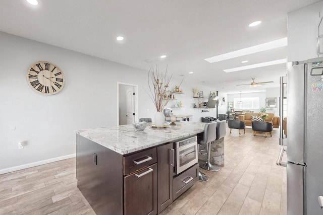 kitchen featuring dark brown cabinetry, light stone counters, a center island, stainless steel appliances, and light wood-type flooring