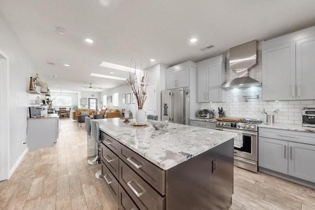 kitchen featuring light wood finished floors, decorative backsplash, appliances with stainless steel finishes, a ceiling fan, and wall chimney range hood